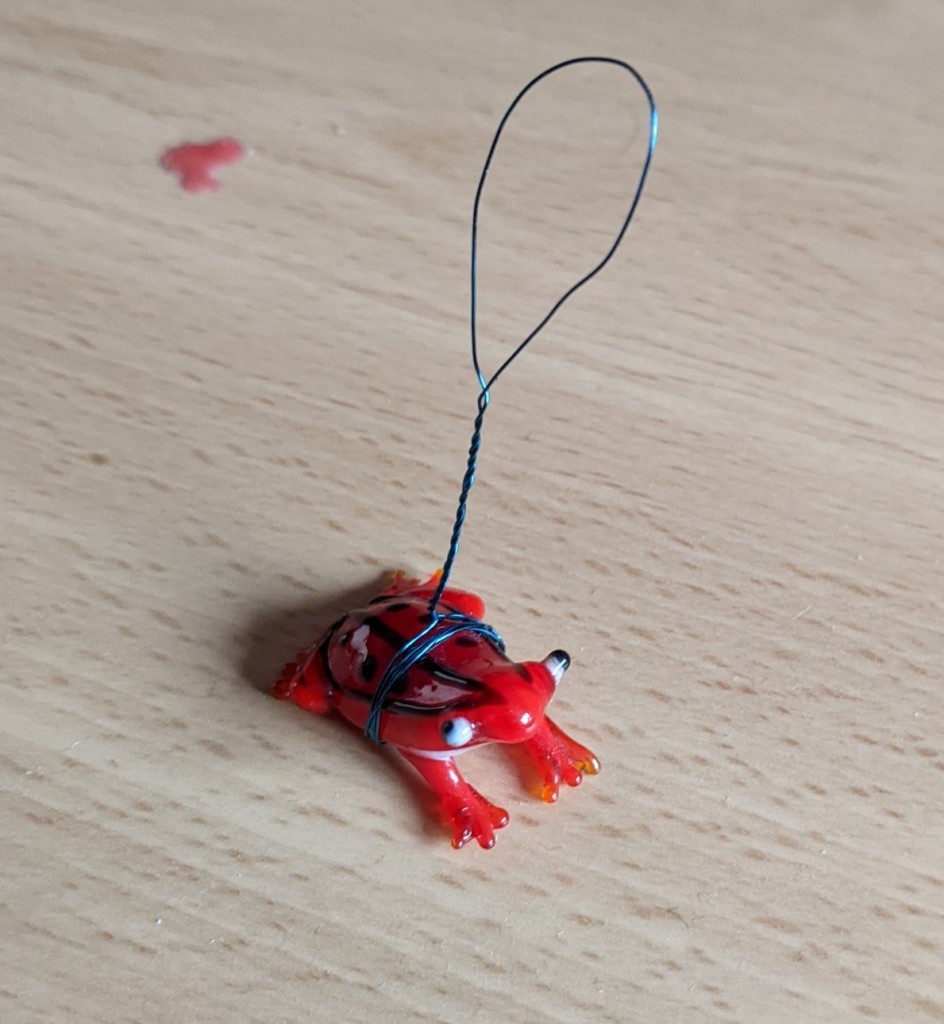 Small frog made of red glass, with a hanging loop of blue wire wrapped around its waist