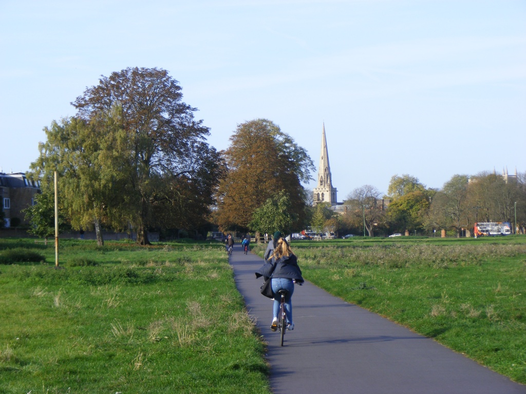 People cycle on a wide path across a grassy common towards a church spire