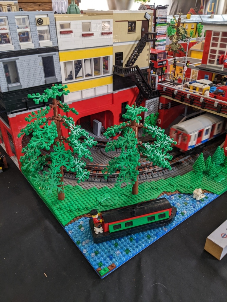 Lego model of buildings, trees, narrowboat on a river, and a London Underground train disappearing into a tunnel under a road.