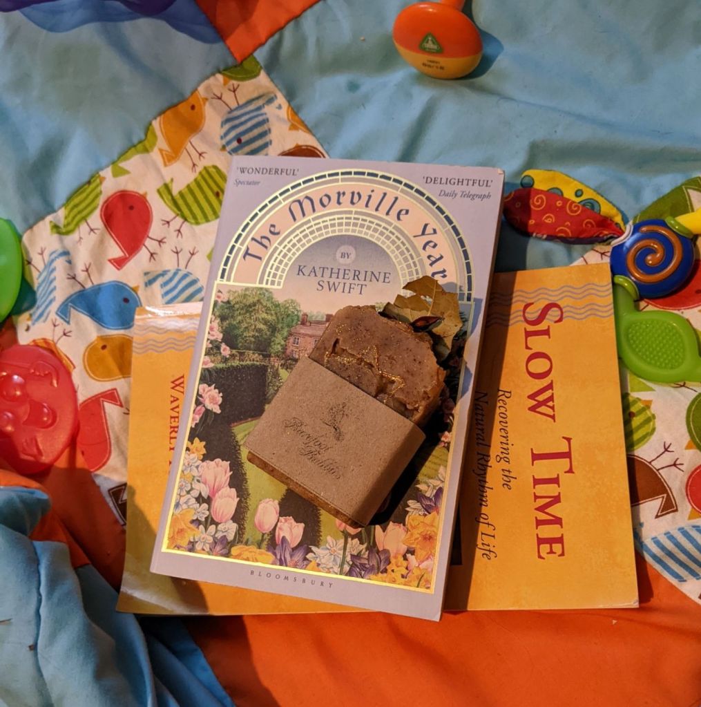 Slow Time by Waverly Fitzgerald, The Morville Year by Katherine Swift, and a bar of soap garnished with star anise and a dried bayleaf, all on a brightly coloured quilt with baby toys 