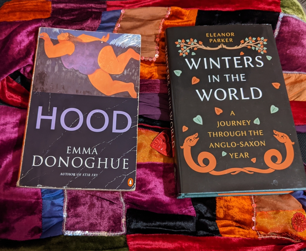 Paperback copy of Hood by Emma Donoghue and a hardback copy of Winters in the World: a journey through the Anglo-Saxon year by Eleanor Parker, both on a brightly coloured velvet patchwork scarf 