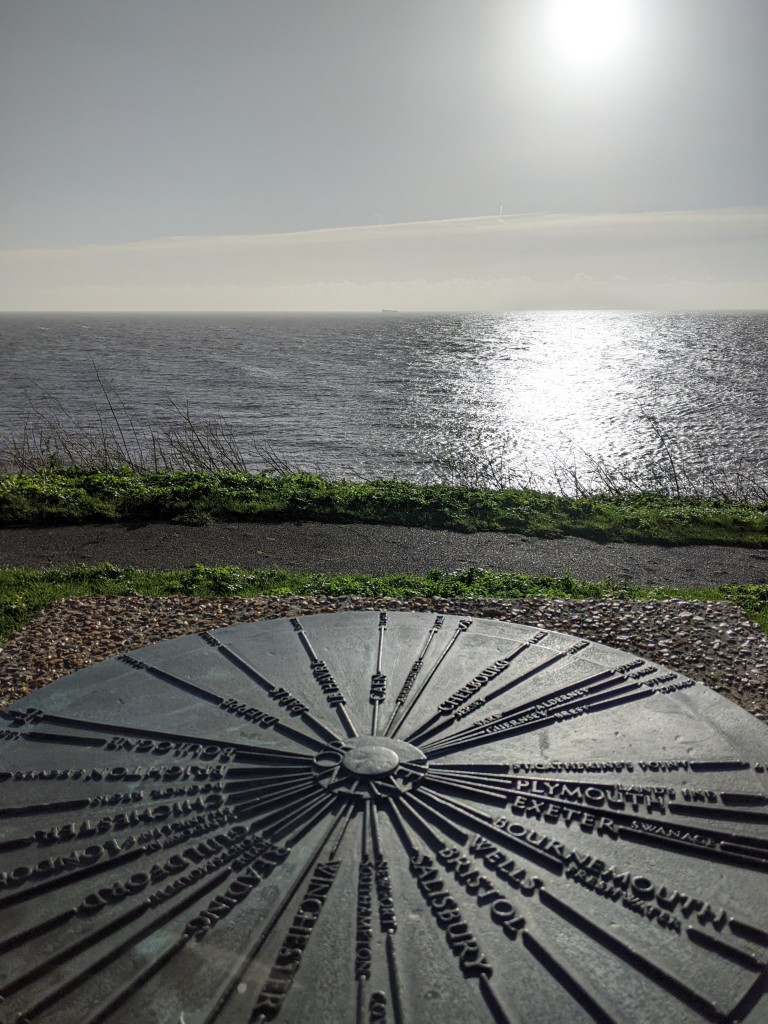 Very bright sun shining on a grey-blue sea. At the front of the shot, a circular diagram gives directions and distances to various towns and cities in England and France. 