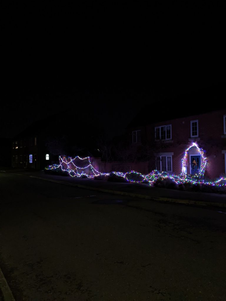 House in darkness, but with the front doorway and hedge strung with multiple strands of coloured fairy lights.