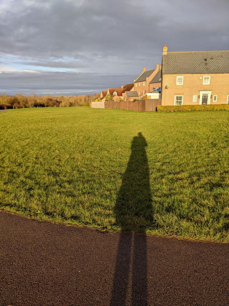 Long winter shadow of a person, rather lumpy, against very bright green grass. The sky beyond is grey layered over blue.