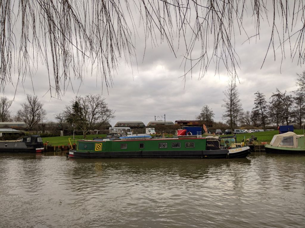 A freight train crosses the back of the picture on a grey day; in front of it, a narrowboat is moored on a river.