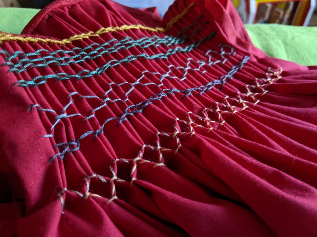 Detail of a child's frock in red fabric with a smocked front. The stitching is somewhat irregular.
